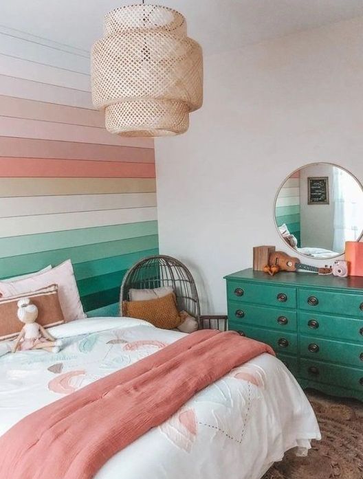 a pretty rainbow girl bedroom with a rainbow accent wall, a bed with pastel and neutral bedding, an emerald dresser with a round mirror