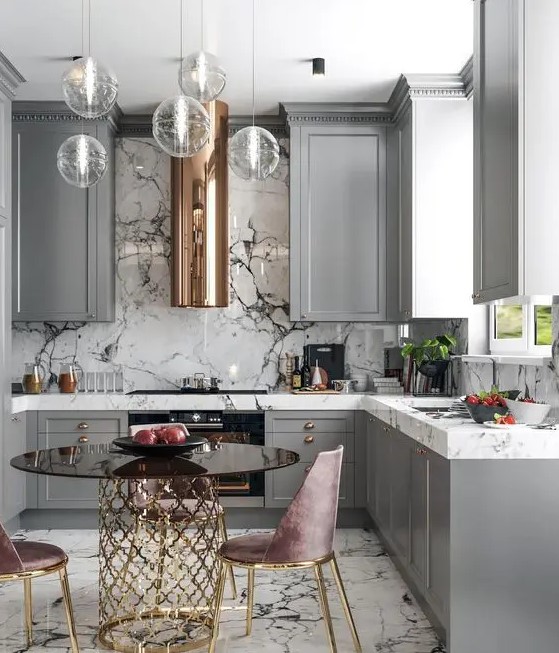 a refined glam ktichen with grey cabinets, white marble coutnertops and a backsplash, a glass table on a gold base, dusty pink chairs and pendant lamps
