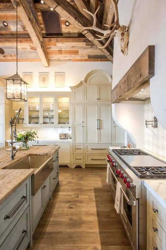 a refined vintage barn kitchen with a wooden ceiling with beams, creamy shaker style cabinets, a light blue kitchen island, butcherblock countertops and pendant lamps