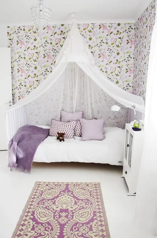 a romantic girl's room with floral wallpaper, a white bed with lilac and white bedding, a white canopy, a white cabinet and a purple printed rug