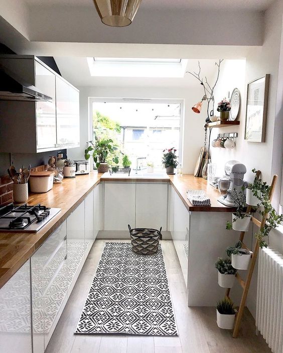 a small white kitchen with butcherblock countertops, potted greenery, a printed rug and pendant lamps