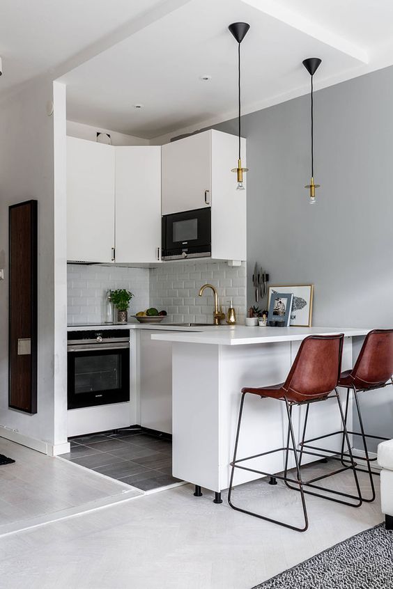 a small white kitchen with white tiles, pendant lamps, brown leather stools and gold touches is chic