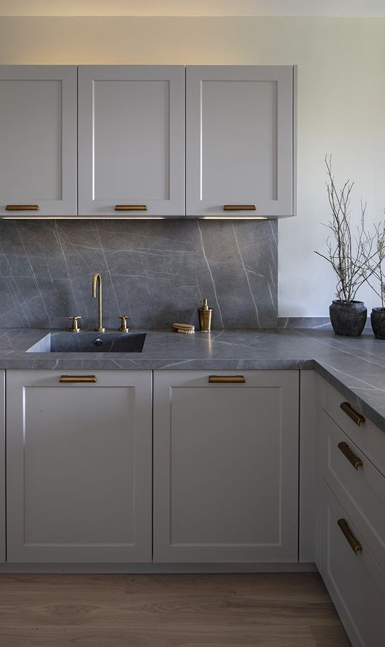 a stylish monochromatic grey kitchen with shaker style cabinets, a grey marble backsplash and countertops plus brass fixtures and handles