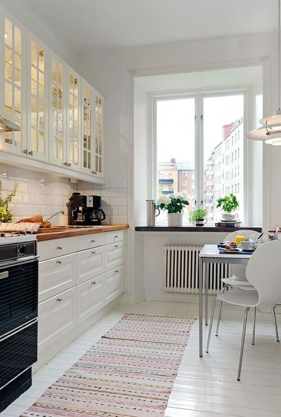 a welcoming white Scandinavian kitchen with glass cabinets, white tiles, butcherblock countertops and a small eating zone