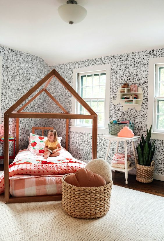 a whimsy girl's room with spotted wallpaper, a house-shaped bed with bright bedding, a white nightstand, a basket with pillows