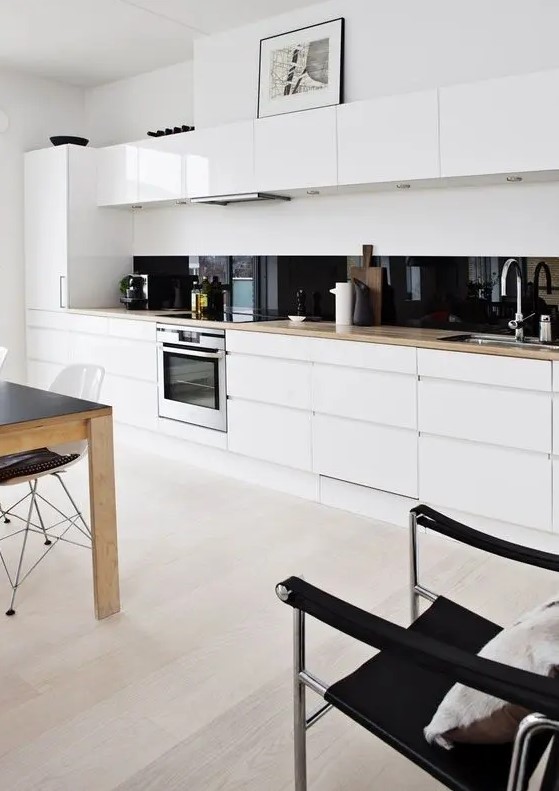 a white minimalist one wall kitchen with blonde butcherblock countertops, a black glass backsplash and an eating zone