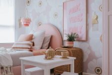 an amazing girl’s bedroom with a floral accent wall, a pink upholstered bed, white play furniture, artwork and a wooden bead chandelier