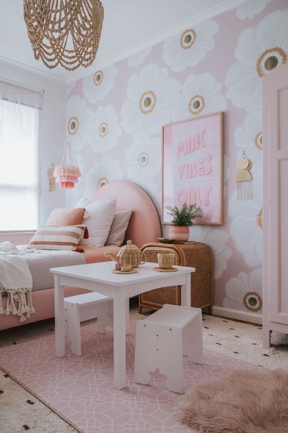 an amazing girl's bedroom with a floral accent wall, a pink upholstered bed, white play furniture, artwork and a wooden bead chandelier