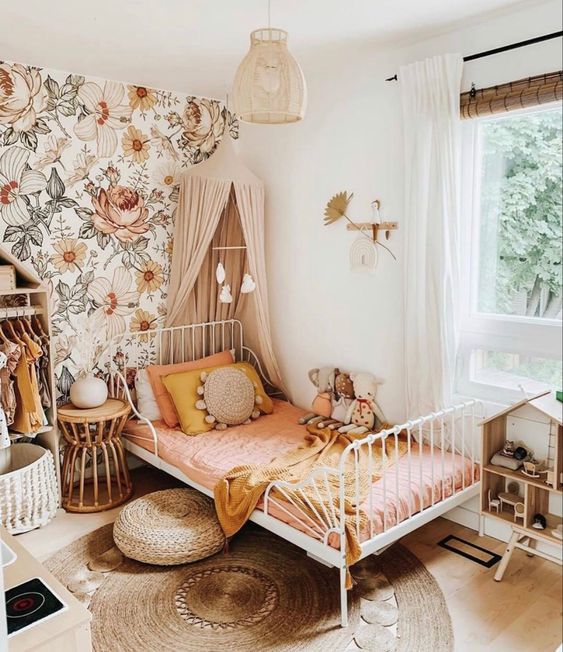 an earthy-tone girl's bedroom with floral wallpaper, a white metal bed with pink bedding, a canopy, an open wardrobe and a play house