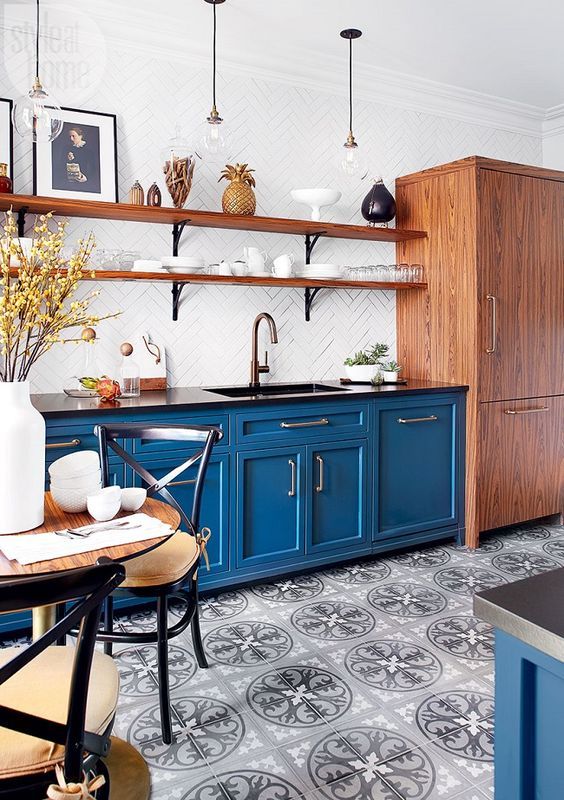 a bold blue kitchen with a printed tile floor, wooden shelves and a storage unit, black chairs with cushions is welcoming