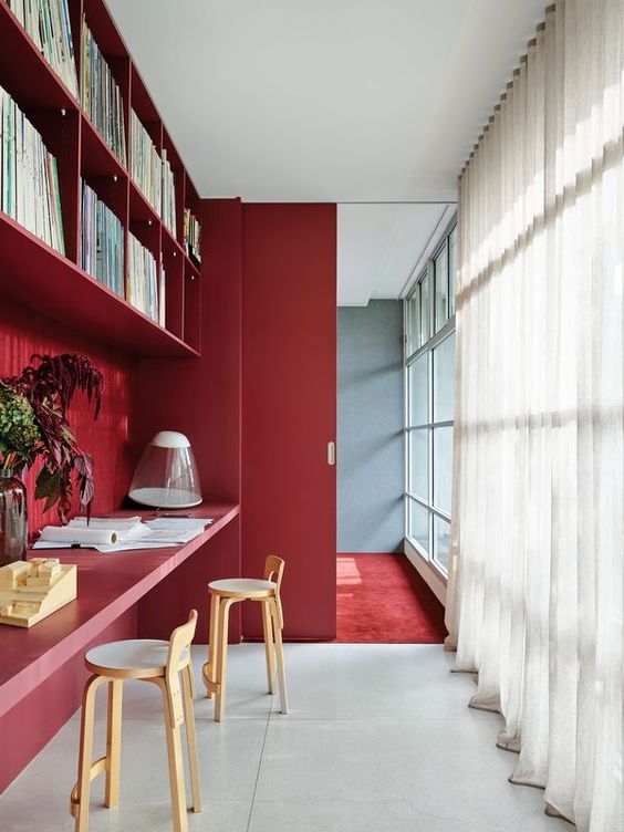 a bold home office with a burgundy accent wall, bookshelves and a built in floating desk, a burgundy sliding door and neutral stools