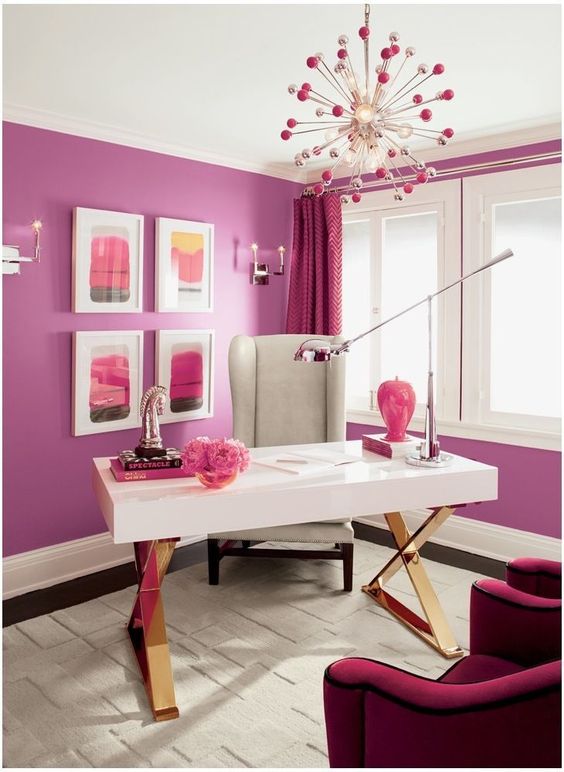 a bright home office with a purple accent wall, an elegant white desk, a grey chair, a fuchsia sofa and a pink and copper burst chandelier