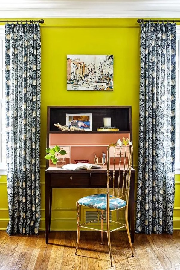 a bright home office with neon yellow walls, a vintage dark stained desk, a gilded chair with a tropical seat and printed curtains