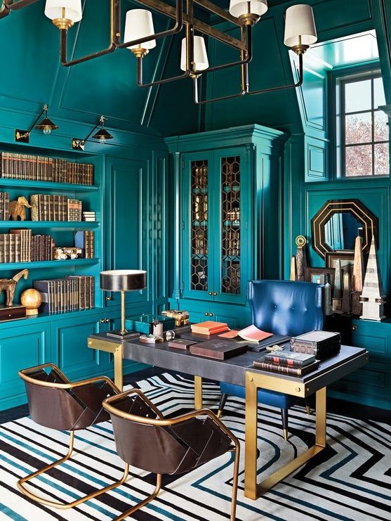 a bright maximalist home office with teal walls and matching built-in furniture and shelves, a refined desk, a navy and leather chairs, a chic chandelier