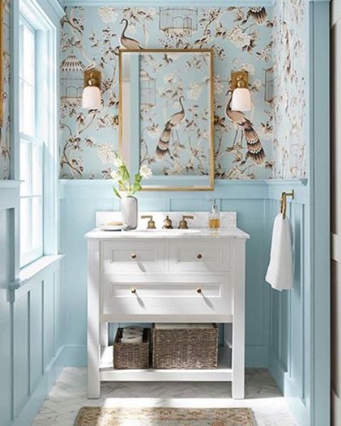 a catchy and airy powder room with light blue panels, beautiful wallpaper, a white vanity, a mirror and sconces