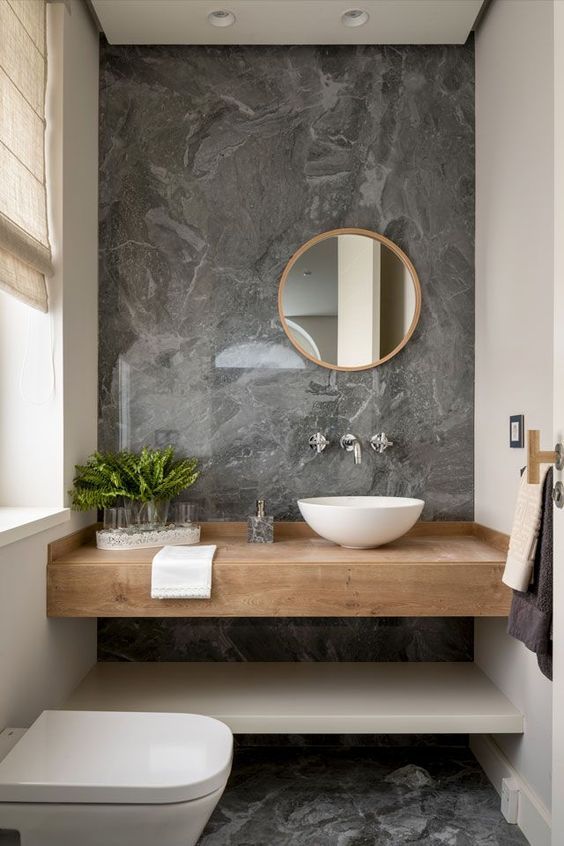 a chic contemporary bathroom with grey marble, a sleek vanity, a bowl sink, a round mirror and a window with a curtain