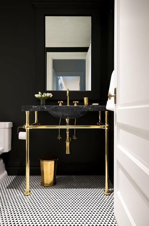 a chic powder room with black walls, a black marble sink, gold fixtures and legs plus a mosaic tile floor