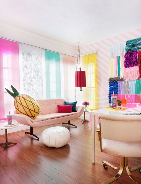 a colorful crafting space with bright curtains hanging, bright pieces of fabric on the wall, a white desk and a chair and a pink sofa plus colorful pillows