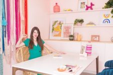 a colorful fancy home office with pink walls, open shelves and a large storage unit, a large desk, a peacock chair, navy chairs and colorful fringe