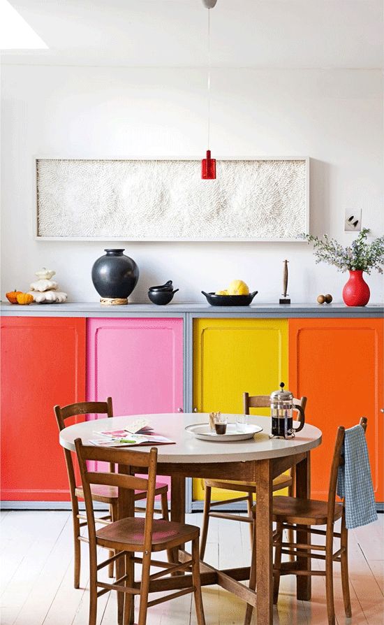 a colorful kitchen with bold doors, and a round table, wooden chairs and a pretty textural artwork and red touches