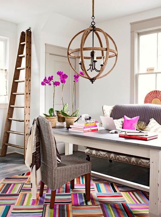 a fancy and whimsy home office with a grey sofa and woven chair, a large desk, a sphere chandelier, a ladder and colorful touches