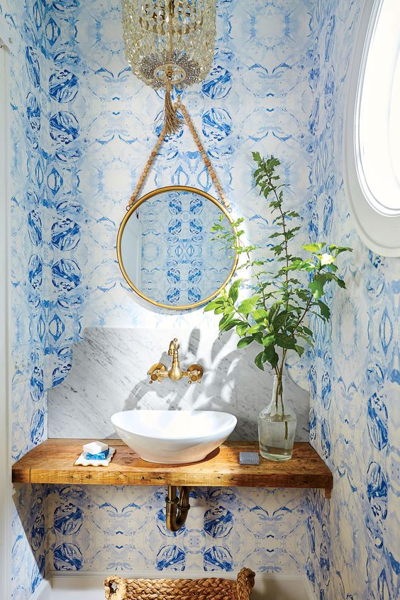 a fantastic powder room with bold blue wallpaper, a wooden vanity, a sink, gold fixtures and a bead chandelier