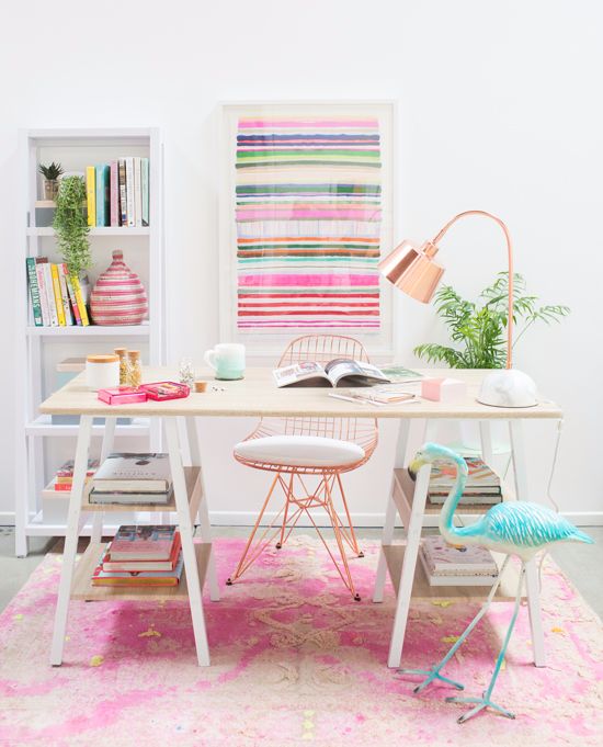 a fun tropical home office with a printed pink rug and a striped artwork, a shelving unit with bright books and a vase, a turquoise flamingo and a copper lamp
