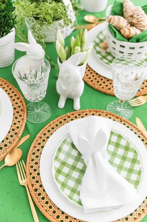 a green and white Easter tablescape with a green tablecloth, rattan placemats, green plaid plates, white tulips, bunny figurines and gold cutlery