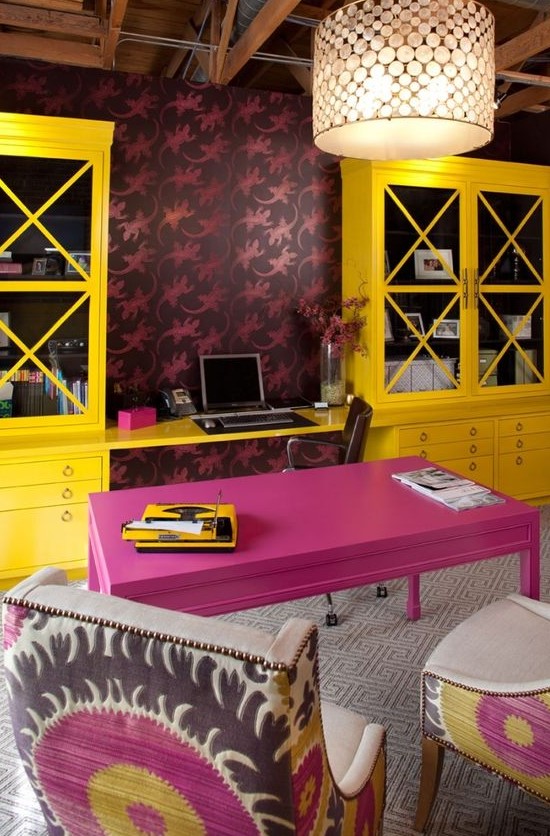 a maximalist home office with bold wallpaper, a hot pink desk and bold yellow storage units, colorful chairs and a pendant lamp