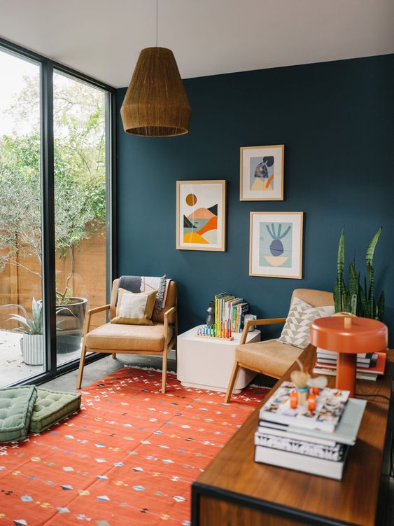 a mid-century modern home office with plenty of color, a dark green accent wall, an orange printed rug, a stained desk and leather chairs, bright artwork and books