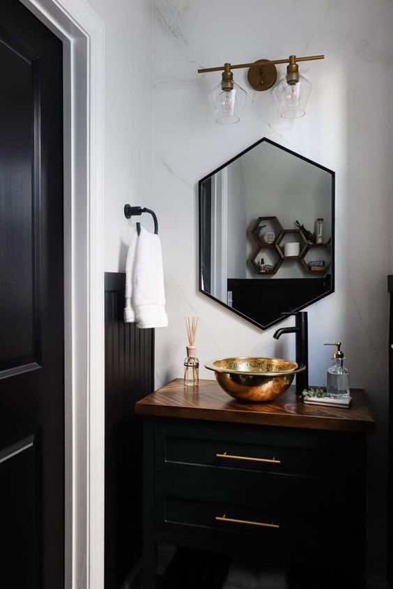 a modern powder room with a black vanity, a hexagon mirror, a gold bowl sink and black fixtures is amazing