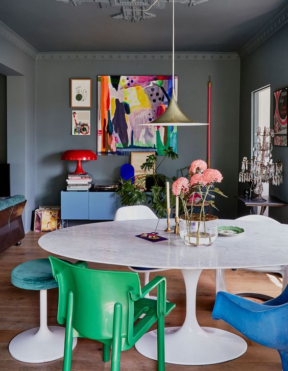 a moody home office made bright with furniture and artwork, with green and blue chairs and stools, a blue dresser, a colorful artwork and a red lamp is a lovely space