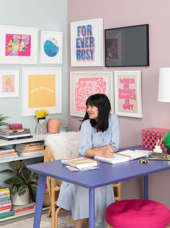 a pastel maximalist home office with a colorful gallery wall, a lilac desk, a hot pink stool and pastel books and a lamp