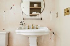 a pretty powder room with pink printed wallpaper, a pedestal sink, a round mirror, sconces, brass touches for a chic touch
