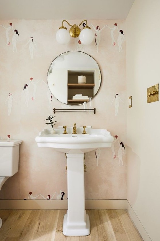 a pretty powder room with pink printed wallpaper, a pedestal sink, a round mirror, sconces, brass touches for a chic touch