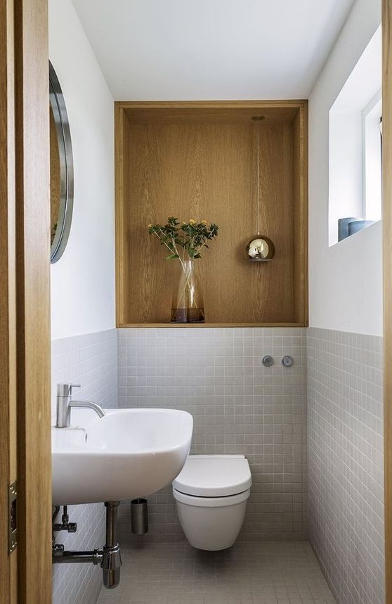 a small powder room with a plywood niche, grey tiles, white appliances including a white floating sink and a round mirror