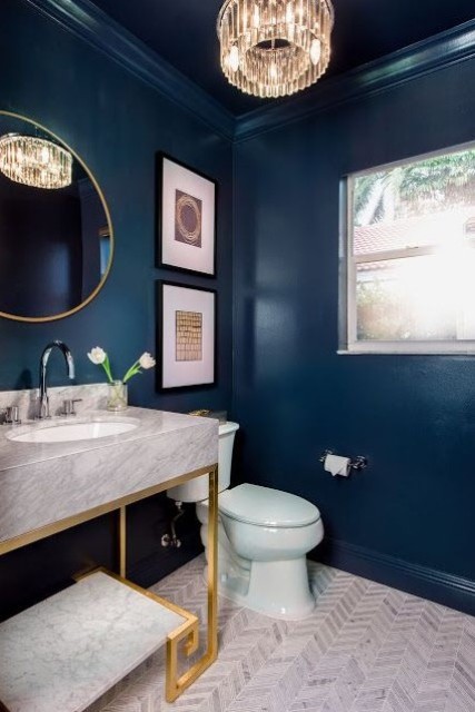 a stylish powder room with navy walls, a crystal chandelier, a gold frame mirror, a gold and white stone vanity and chic artworks