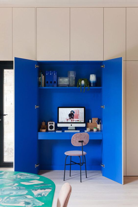 a super colorful hidden home office nook done in electric blue, with a built in shelf and a small desk with drawers, a blush chair