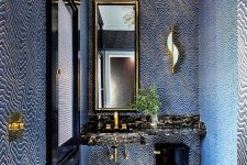 a very quirky powder room with navy and gold pritned wallpaper, a black stone vanity and a black toilet plus a unique chandelier