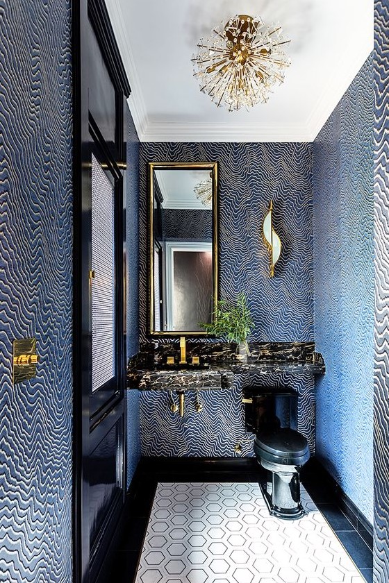 a very quirky powder room with navy and gold pritned wallpaper, a black stone vanity and a black toilet plus a unique chandelier