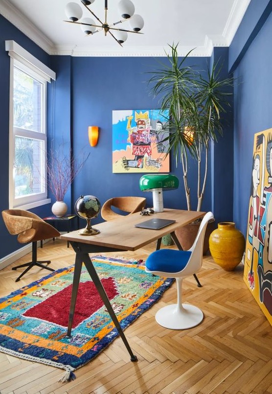 a vibrant home office with bold blue walls, colorful artworks and a rug, mismatching chairs and a statement plant in the corner