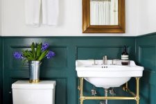 an elegant powder room with teal panels on the walls, a console sink, a mirror in a brass frame, white appliances and neutral towels