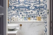an eye-catchy powder room with blue wallpaper, white appliances, white marble tiles, gold touches for more glam