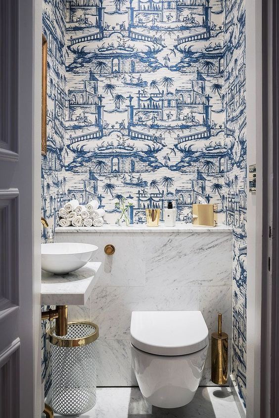 an eye-catchy powder room with blue wallpaper, white appliances, white marble tiles, gold touches for more glam