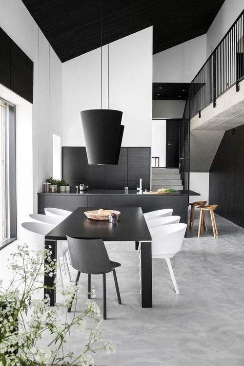 a minimalist black and white dining room with a black table, white curved chairs, black pendant lamps and a black kitchen next to it