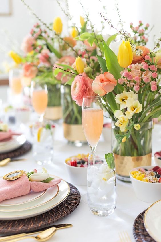 a bright Mother's Day tablescape with yellow and pink blooms, greenery and twigs, wicker placemats, pink napkins and gold cutlery