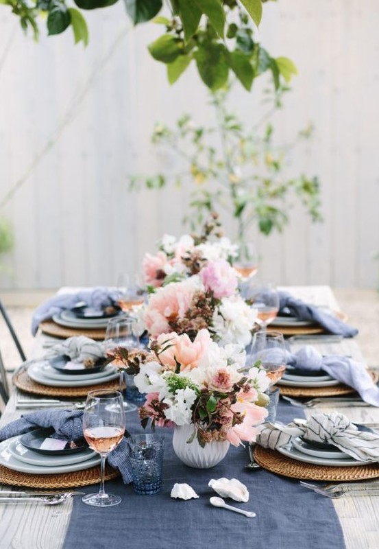 a bright spring table setting with navy napkins and a table runner, wicker chargers and blue porcelain and pink and white floral centerpieces