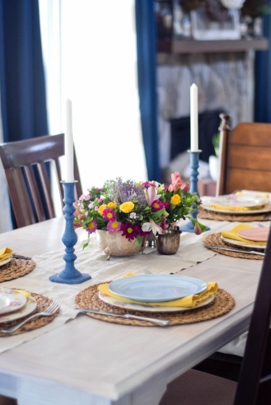 a colorful spring table setting with a bright floral centerpiece, wicker chargers, blue candle holders, yellow napkins