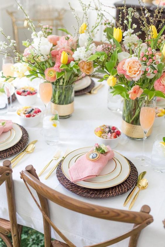 a lively Mother's Day tablescape with coral and yellow blooms and blooming branches, wicker placemats, pink napkins and gold cutlery