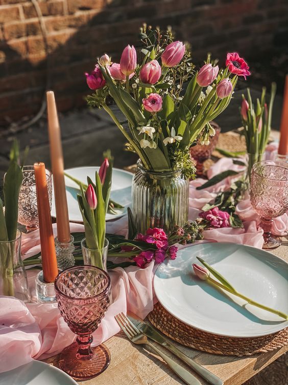 a sophisticated pink Mother's Day tablescape with a pink runner, pink blooms and glasses, ornage candles and wicker placemats